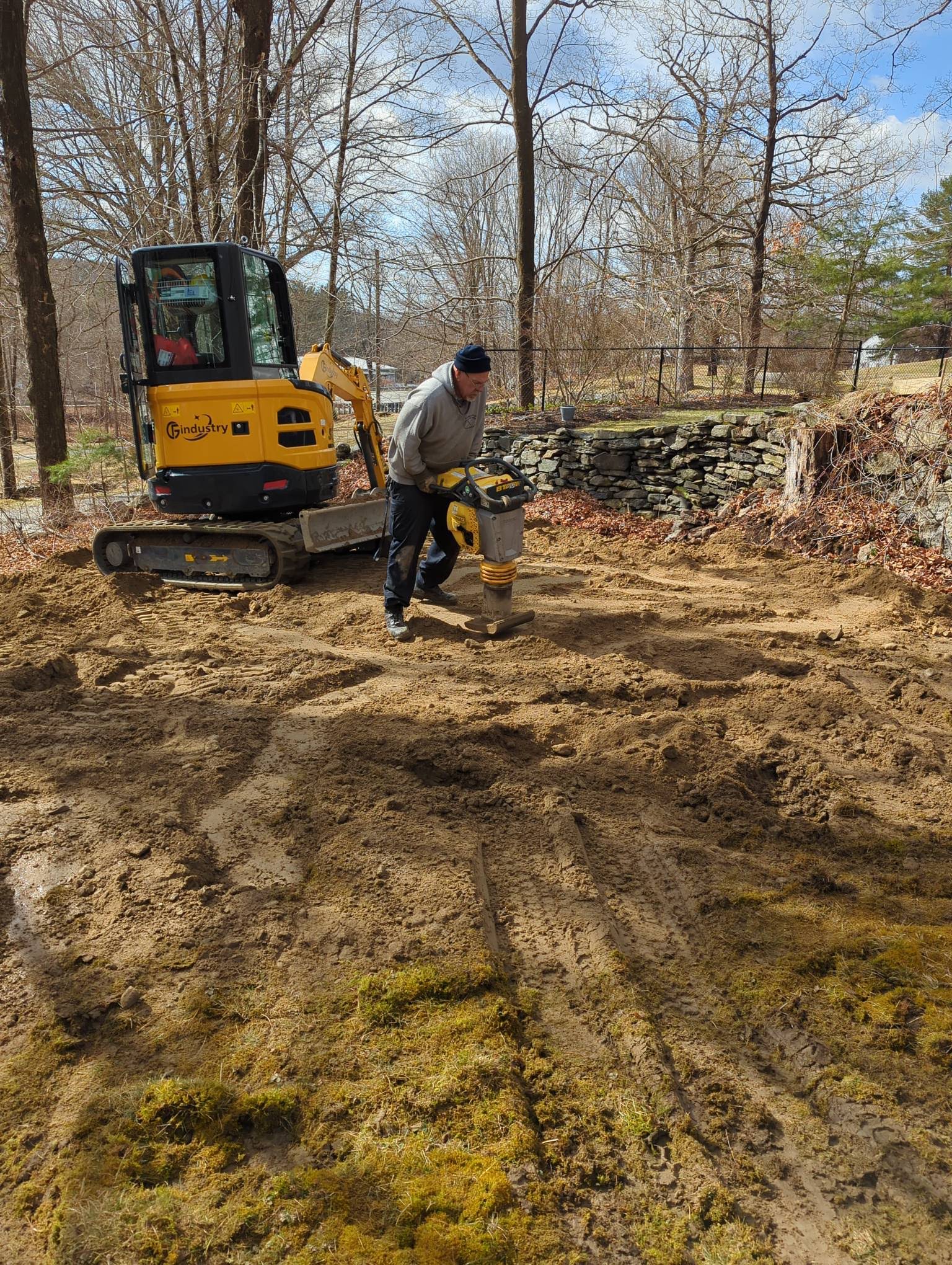 Worker operating plate compactor with excavator near stone wall