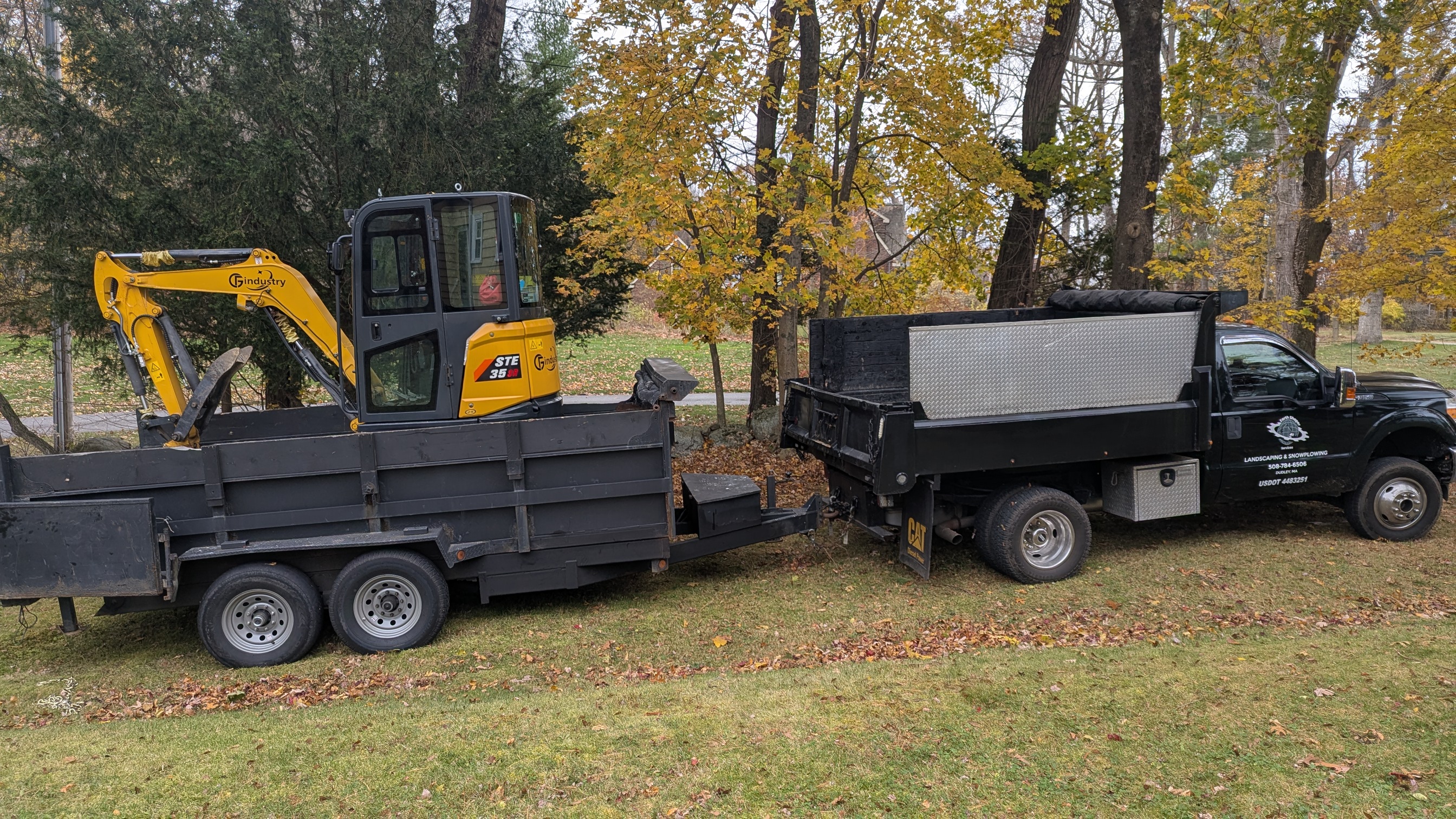 Yellow Industry excavator loaded on trailer next to Earth Shaper Services dump truck during autumn